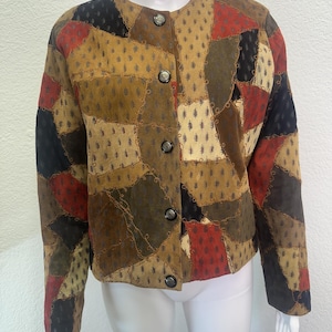 May include: A patchwork jacket in shades of brown, tan, red, and black. The jacket features a round collar, long sleeves, and four dark buttons down the front. The fabric has a textured, suede-like appearance, with a variety of stitched patches.
