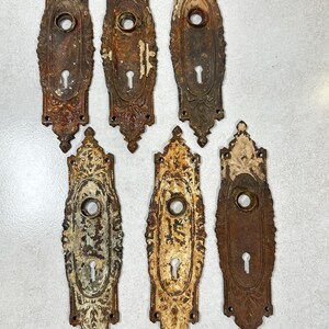 May include: Six elongated, rectangular metal door plates with pointed ends, featuring a central circular opening for a knob and a lower keyhole. The plates exhibit varying degrees of weathered, rusted brown and chipped off-white paint finishes.