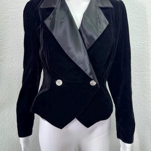 May include: Black velvet blazer with a satin lapel and two decorative silver buttons. The blazer has a tailored fit and long sleeves. The jacket is displayed on a mannequin.