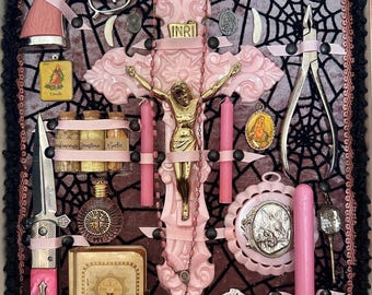 Può includere: Una collezione di oggetti religiosi e gotici rosa e argento disposti in una scatola ombra. Include un crocifisso, una pistola giocattolo, candele, un coltello e altri elementi decorativi. Lo sfondo presenta un disegno a ragnatela nera.