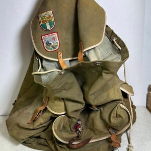 May include: An olive green canvas backpack with multiple pockets and leather straps. The backpack features two patches, one with the word "Rotterdam" and the other with "Luxembourg". The bag has a drawstring closure and a white trim.