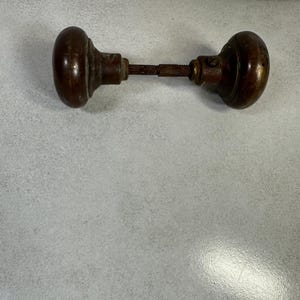 May include: A pair of dark brown, spherical wooden door knobs connected by a central metal spindle. The knobs feature a smooth, rounded surface with visible signs of wear and aging, including a small metal screw visible on the right knob.