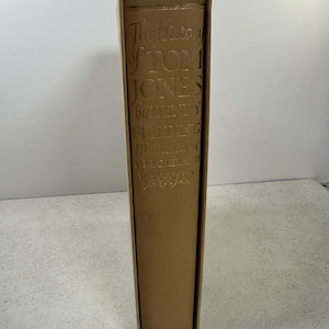 May include: A tan hardcover book titled The History of Tom Jones by Henry Fielding, illustrated by T.M. Cleland. The spine features gold embossed lettering and decorative flourishes, housed within a matching tan slipcase with a vertical orientation.