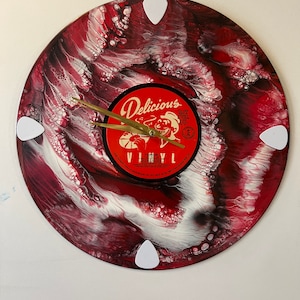 May include: A red, white, and black vinyl record clock with a gold clock hand. The record has a red label with the text "Delicious Vinyl" and a cartoon character. The clock is decorated with four white guitar picks.