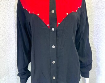 May include: A long-sleeved button-down shirt featuring a black body and a vibrant red western-style yoke. The shirt includes a pointed collar adorned with small silver studs, matching silver studs along the yoke, and decorative silver buttons.