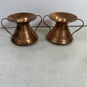 May include: A pair of matching copper vessels with wide, flared rims and two thin, twisted metal handles on each side. The bodies of the vessels feature a conical shape with subtle horizontal etched lines around the lower section and a polished finish.