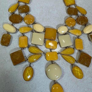 May include: A statement necklace featuring a variety of geometric shapes in shades of yellow, cream, and brown. The necklace has a silver-tone chain and a mix of oval, square, and teardrop-shaped pendants.