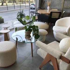 Op de afbeelding: Een woonkamer met witte, pluche meubels, waaronder een grote fauteuil, een kleinere fauteuil en verschillende ronde poefs. Een glazen salontafel met een houten onderstel staat in het midden van de kamer, omringd door de meubels. Een plant in een pot met groene bladeren voegt een vleugje natuur toe aan de ruimte.