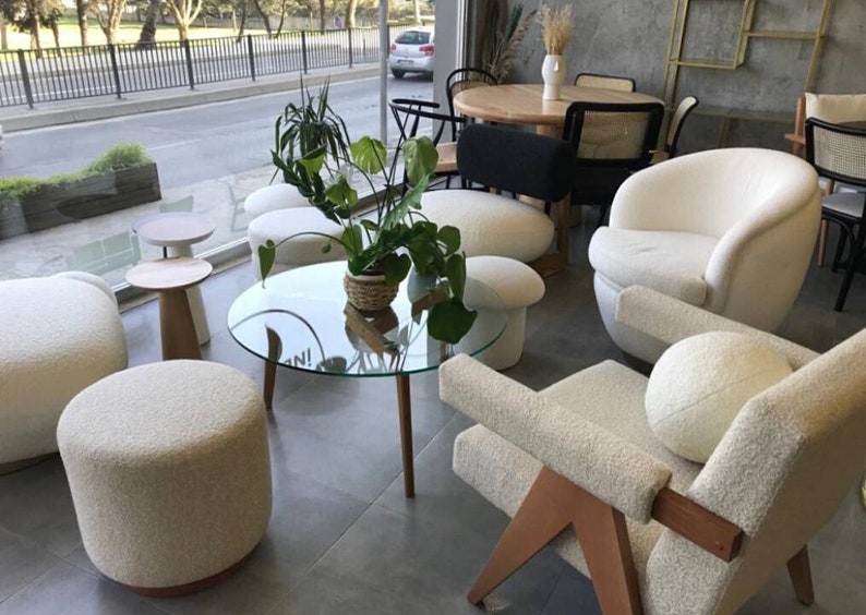 Op de afbeelding: Een woonkamer met witte, pluche meubels, waaronder een grote fauteuil, een kleinere fauteuil en verschillende ronde poefs. Een glazen salontafel met een houten onderstel staat in het midden van de kamer, omringd door de meubels. Een plant in een pot met groene bladeren voegt een vleugje natuur toe aan de ruimte.