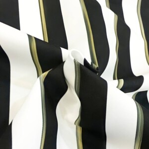 May include: Close-up of a fabric with a bold striped pattern. The design features alternating vertical stripes of black and white, with thin olive green stripes. The fabric is folded, creating depth and texture.