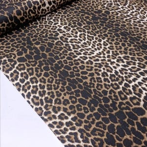 May include: A bolt of fabric featuring a repeating leopard print pattern. The design consists of dark black spots with tan and brown centers arranged in a dense, organic layout across the surface of the textile, which is folded at the top edge.