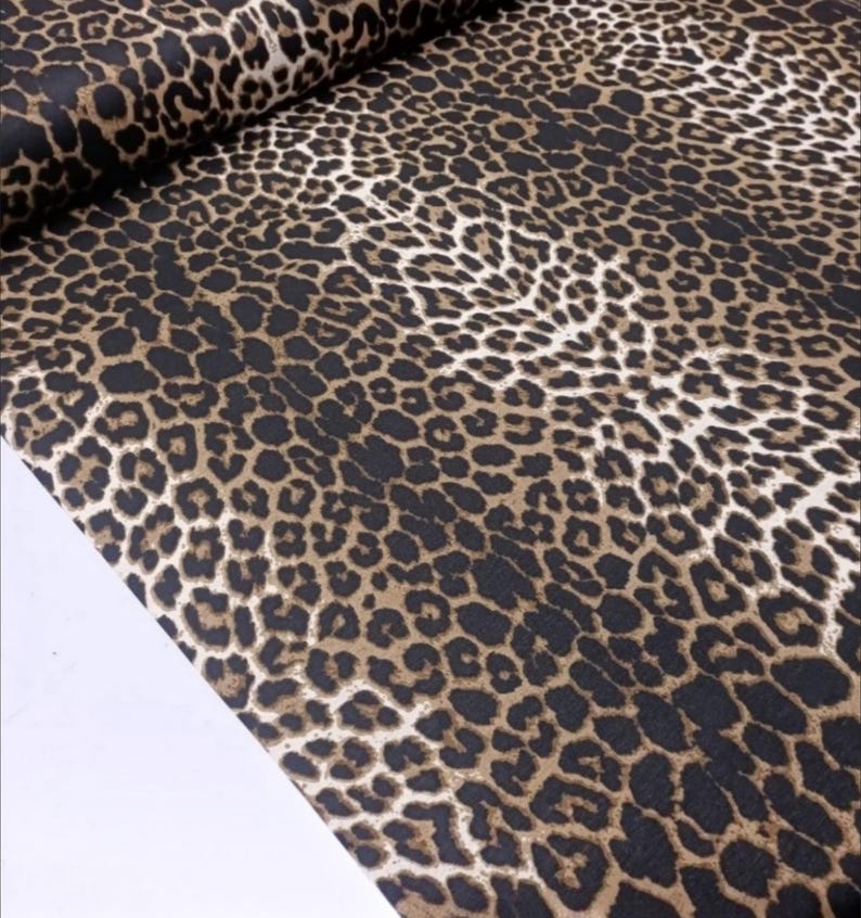 May include: A bolt of fabric featuring a repeating leopard print pattern. The design consists of dark black spots with tan and brown centers arranged in a dense, organic layout across the surface of the textile, which is folded at the top edge.
