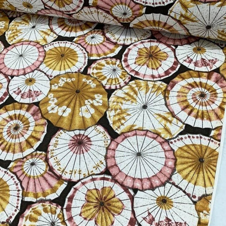 May include: A black fabric with a repeating pattern of white and gold umbrellas with pink accents. The umbrellas are arranged in a random pattern.