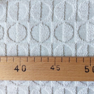 May include: Close-up of a white fabric with a textured pattern of circles and vertical lines. A wooden ruler with markings for 40, 45, and 50 is placed on the fabric.