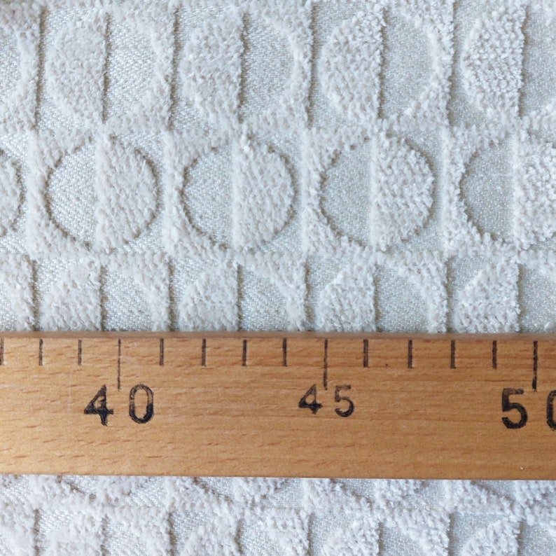 May include: Close-up of a white fabric with a textured pattern of circles and vertical lines. A wooden ruler with markings for 40, 45, and 50 is placed on the fabric.