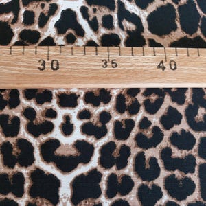May include: A fabric featuring a repeating leopard print pattern in shades of black, brown, and tan. A wooden ruler is placed horizontally across the fabric, showing markings from 30 to 40 centimeters to indicate the scale of the print design.