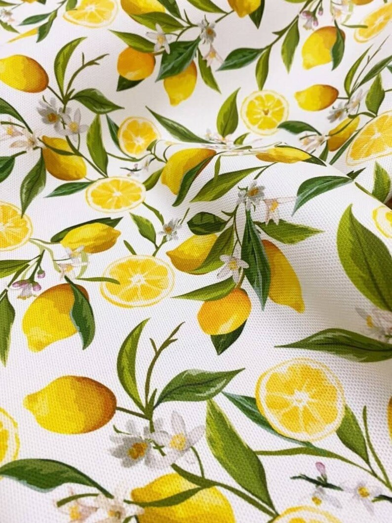 K&ouml;nnte beinhalten: Wei&szlig;er Stoff mit einem sich wiederholenden Muster aus gelben Zitronen, gr&uuml;nen Bl&auml;ttern und wei&szlig;en Bl&uuml;ten. Einige Zitronen sind ganz, andere sind halbiert. Das Design erinnert an einen Sommergarten.