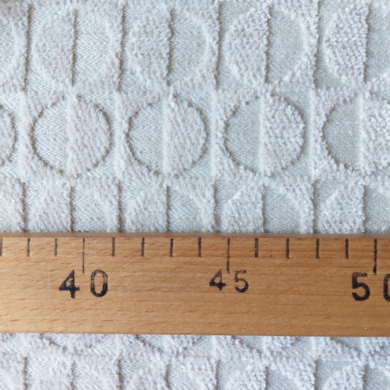 May include: Close-up of a textured, off-white fabric with a repeating circular pattern. A wooden ruler with black markings and numbers is placed across the bottom, measuring from 40 to 50 inches.