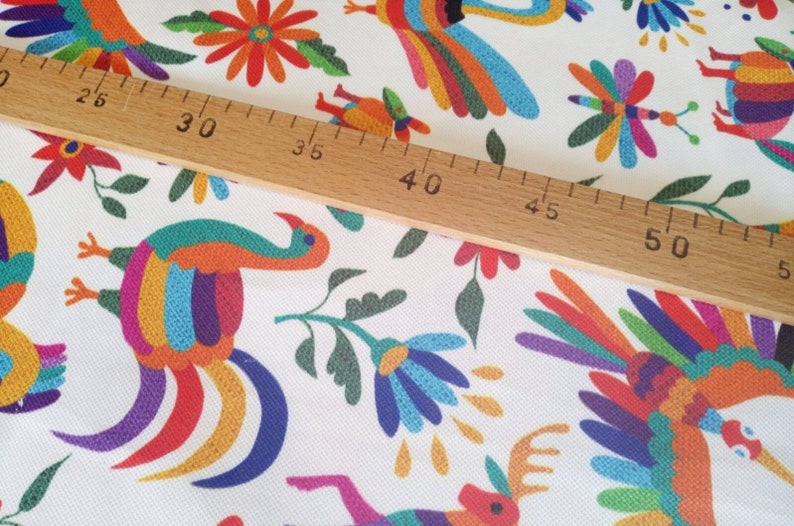 May include: White fabric with a colorful repeating pattern of stylized birds, flowers, and geometric shapes. A wooden ruler is placed on top of the fabric, with the numbers 25, 30, 35, 40, 45, 50, and 55 visible.