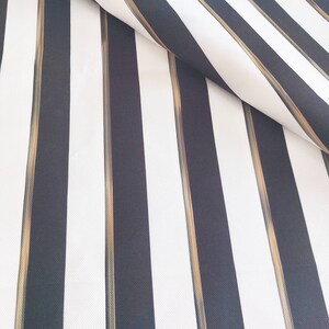 May include: Close-up of a fabric with a bold striped pattern. The design features alternating vertical stripes in black and white, with thin gold-colored stripes running between the black stripes. The fabric is folded over, revealing its texture.