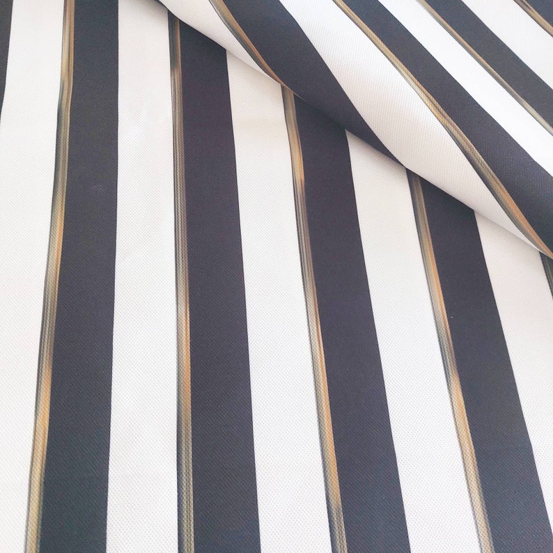 May include: Close-up of a fabric with a bold striped pattern. The design features alternating vertical stripes in black and white, with thin gold-colored stripes running between the black stripes. The fabric is folded over, revealing its texture.
