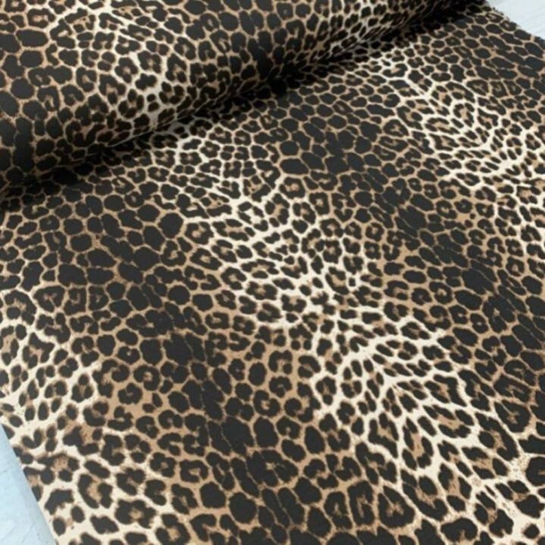 May include: A bolt of fabric featuring a repeating leopard print pattern. The design consists of dark brown and black spots arranged in irregular clusters against a light tan background, creating a dense, continuous animal skin motif across the surface.