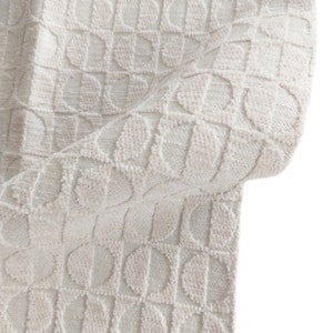 May include: Close-up of a cream-colored fabric with a repeating geometric pattern of half-circles within squares. The fabric has a textured, plush appearance, with a slight sheen. The fabric is folded, revealing its thickness and construction.