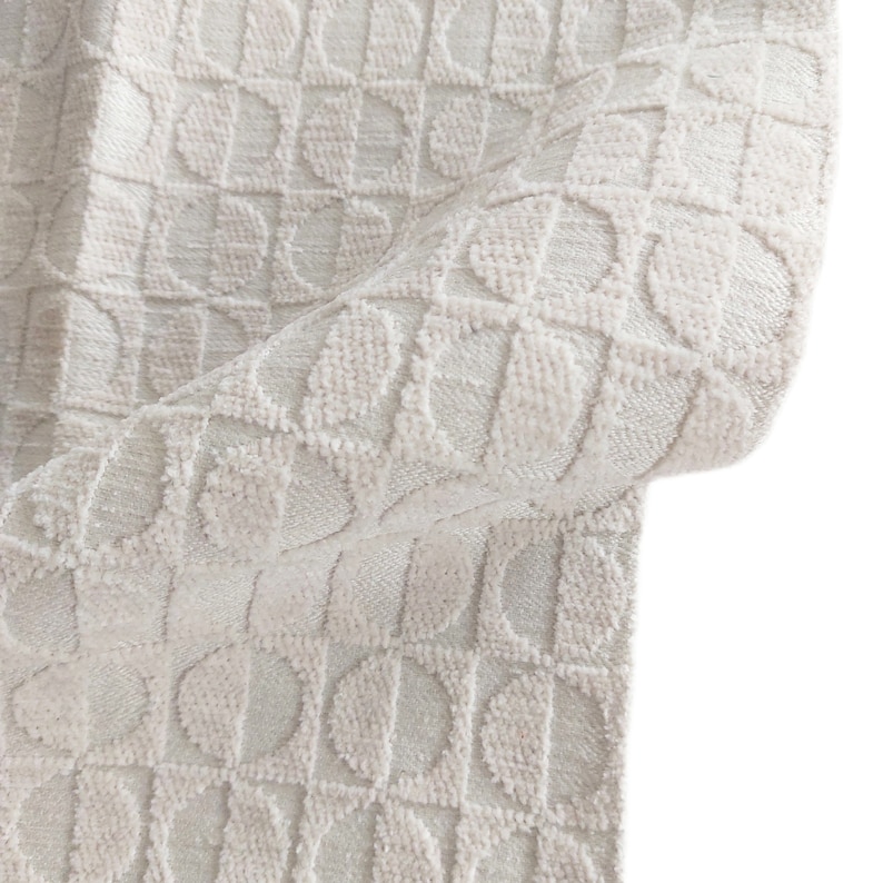 May include: Close-up of a cream-colored fabric with a repeating geometric pattern of half-circles within squares. The fabric has a textured, plush appearance, with a slight sheen. The fabric is folded, revealing its thickness and construction.