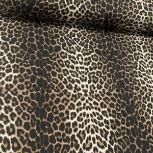 May include: A fabric featuring a repeating leopard print pattern with black spots outlined in dark brown against a tan background. The material is folded at the top, showing a consistent texture and a dense, detailed animal skin design.