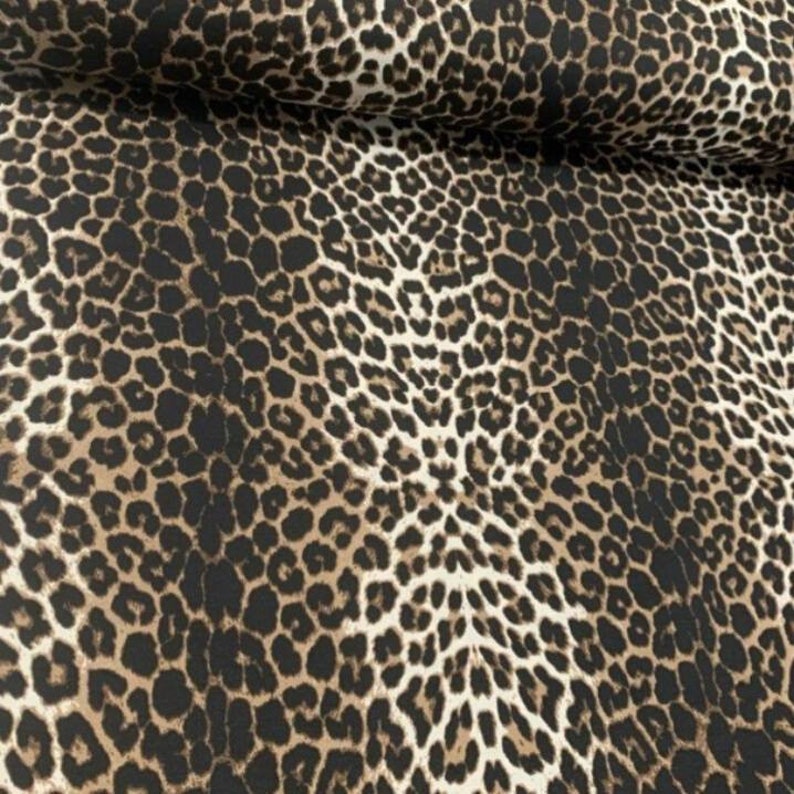 May include: A fabric featuring a repeating leopard print pattern with black spots outlined in dark brown against a tan background. The material is folded at the top, showing a consistent texture and a dense, detailed animal skin design.