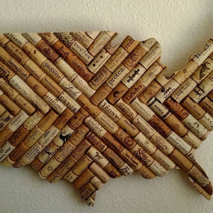 May include: A decorative wall hanging in the shape of the United States, crafted from numerous wine corks. The corks are arranged in a diagonal pattern, showcasing various brand names and logos. The overall color is a warm, natural brown.