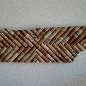 Może przedstawiać: Dekoracja ścienna w kształcie stanu Tennessee, wykonana z przetworzonych kork&oacute;w do wina. Korki ułożone są w jodełkę, prezentując mieszankę jasnych i ciemnych odcieni brązu. Element jest zamontowany na białym tle.