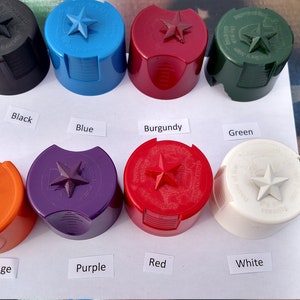 Może przedstawiać: Kolekcja kolorowych kork&oacute;w do butelek w kolorach czarnym, niebieskim, bordowym, zielonym, pomarańczowym, fioletowym, czerwonym i białym. Każdy korek ma gwiazdę na g&oacute;rze i jest oznaczony kolorem. Korki są ułożone na białej powierzchni.