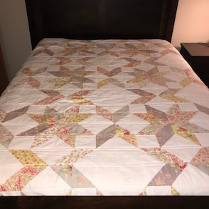 May include: A white quilt with a star pattern in shades of pink, yellow, and gray. The quilt is made of fabric with a floral print.