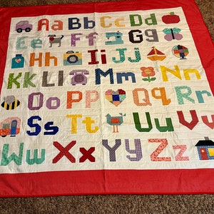 May include: A white alphabet quilt with a red border. Each letter is a different color and has a small picture next to it. The letters are A-Z.