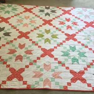 May include: A white quilt with a repeating pattern of red, green, and pink stars and crosses. The quilt is made with different patterned fabrics and has a red border.