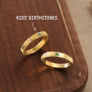 May include: Two gold-colored rings with colorful gemstones are displayed on a wooden surface. The rings are adorned with various birthstones, and the text "KIDS' BIRTHSTONES" is visible above them. The rings are a delicate and elegant jewelry item.