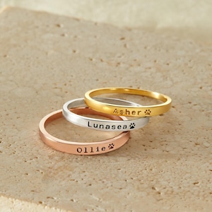 Könnte beinhalten: Drei personalisierte Ringe mit eingravierten Pfotenabdrücken. Die Ringe sind übereinander gestapelt. Der obere Ring ist golden, der mittlere Ring ist silberfarben und der untere Ring ist roségoldfarben. Die Namen "Ascher", "Lunasea" und "Ollie" sind in die Ringe graviert.
