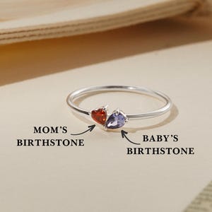 May include: A silver ring featuring two teardrop-shaped gemstones, one red and one purple. Text indicates the red stone is the "MOM'S BIRTHSTONE" and the purple stone is the "BABY'S BIRTHSTONE". The ring is displayed on a neutral background.