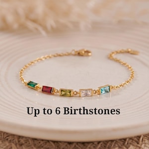 May include: A gold-toned chain bracelet featuring five rectangular baguette-cut gemstones in green, red, yellow, clear, and blue. The text "Up to 6 Birthstones" is printed below the bracelet. The jewelry has a delicate link design and a clasp.