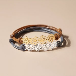 May include: Two bracelets with floral designs. One bracelet has a gold-colored floral design and brown cord. The other bracelet has a silver-colored floral design and gray cord. Both bracelets are on a neutral background.