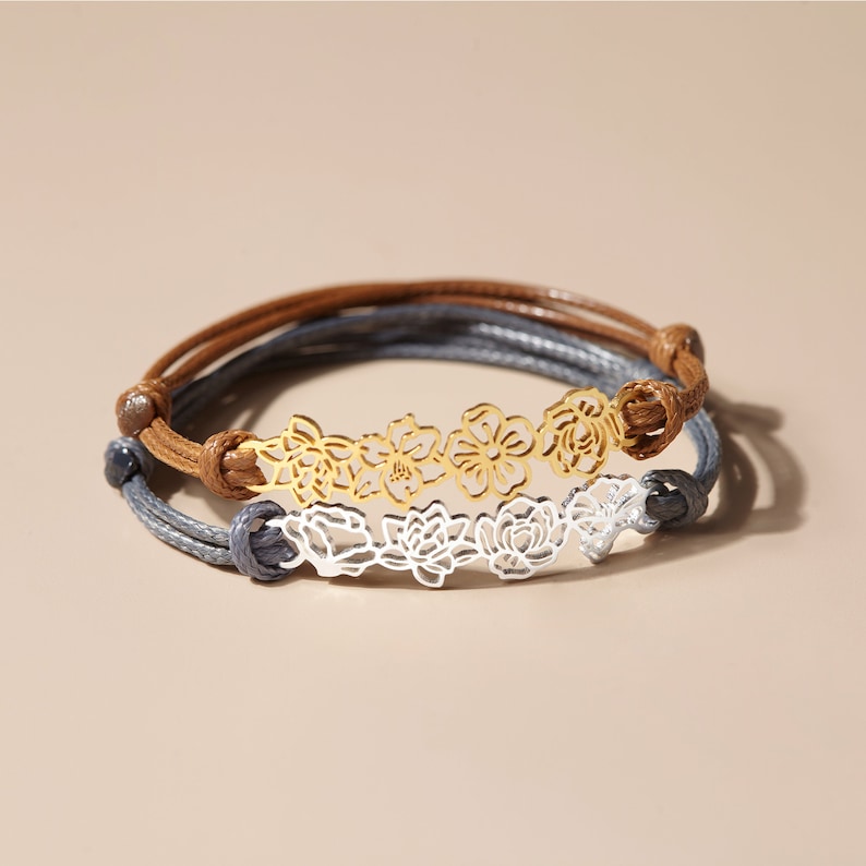 May include: Two bracelets with floral designs. One bracelet has a gold-colored floral design and brown cord. The other bracelet has a silver-colored floral design and gray cord. Both bracelets are on a neutral background.