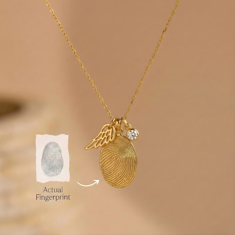 May include: A gold-toned necklace featuring an oval pendant engraved with a fingerprint, a small wing charm, and a round clear gemstone charm. A small white card with a gray fingerprint illustration and the text Actual Fingerprint is included.