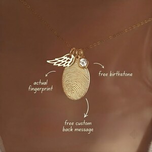 May include: A gold-toned necklace featuring an oval pendant engraved with an actual fingerprint, a small angel wing charm, and a clear round birthstone. Text on the image reads: actual fingerprint, free birthstone, and free custom back message.