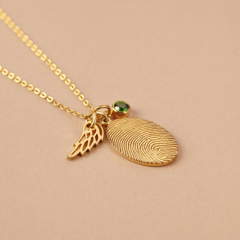 A stunning gold necklace featuring a fingerprint charm, angel wing, and green birthstone on a simple beige background for a clean, classy look.
