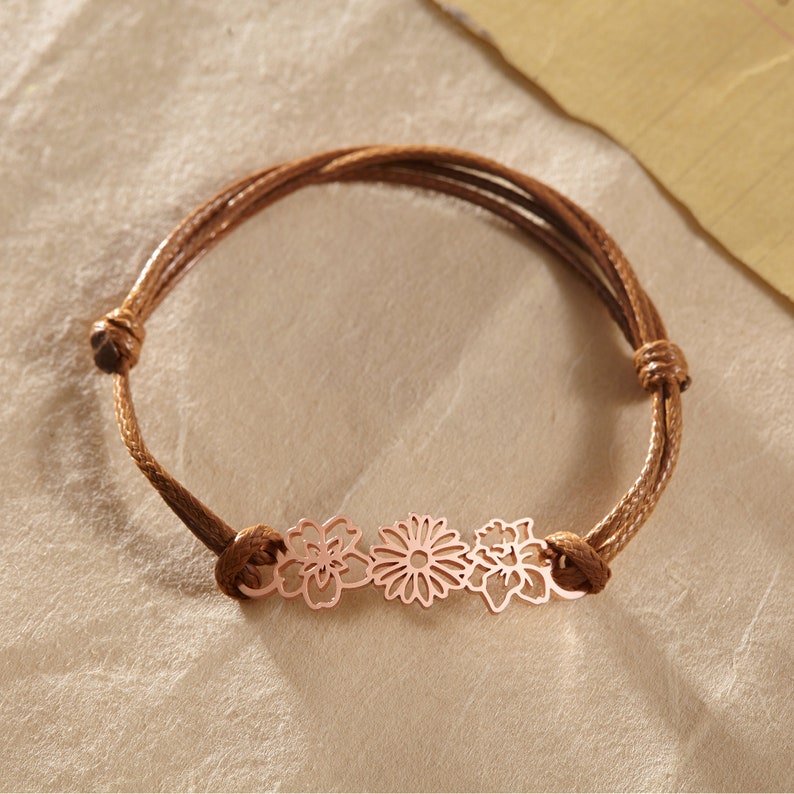 May include: A brown cord bracelet featuring three rose gold flower charms. The bracelet has an adjustable knot closure. The flowers are a daisy, a lotus, and a stylized flower. The bracelet is on a neutral background.