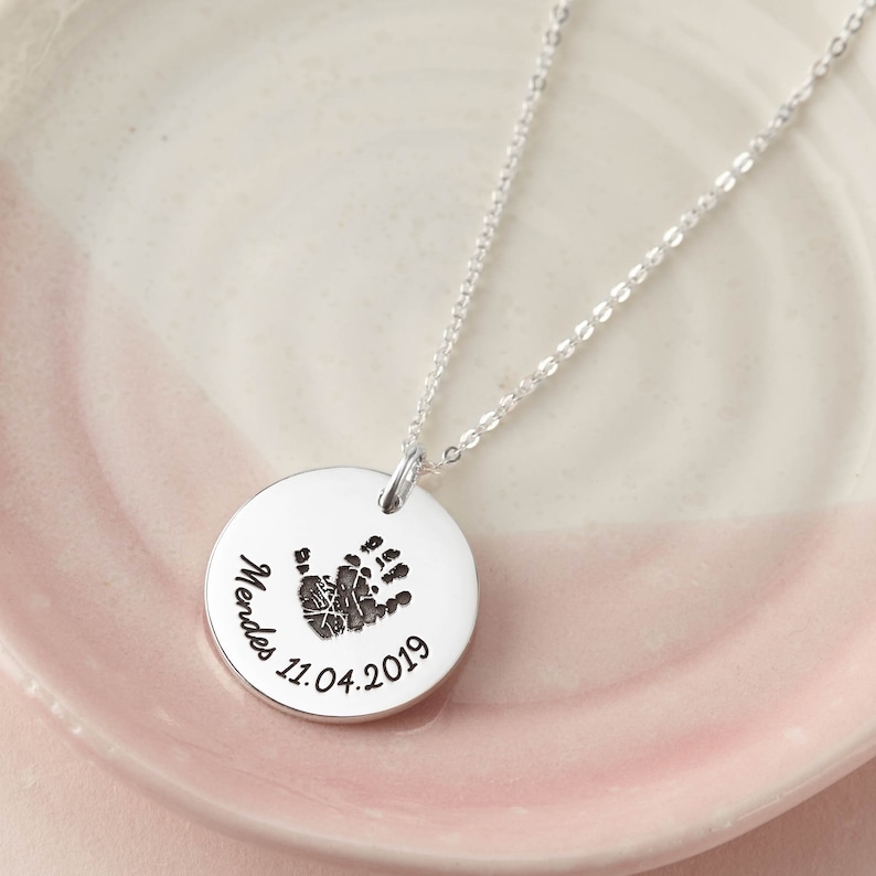 May include: A silver-toned circular pendant necklace featuring an engraved dark handprint at the center. The pendant is inscribed with the name Mendes and the date 11.04.2019 in a black script font. The piece includes a delicate silver-toned chain.