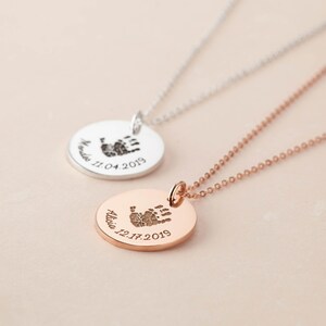 May include: Two circular pendant necklaces, one in silver and one in rose gold. Each pendant features an engraved handprint at the center, with a name and date inscribed along the bottom edge. The silver pendant reads "Marides 11.04.2019" and the rose gold pendant reads "Alicia 12.17.2019".