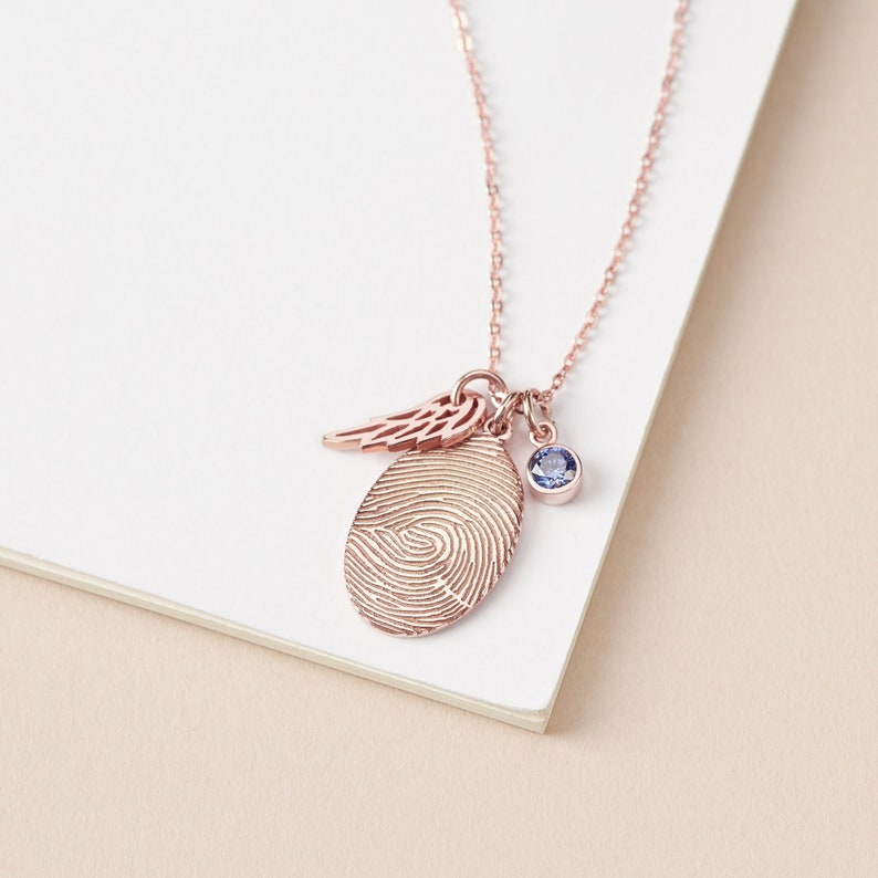 A delicate necklace featuring a personalized fingerprint charm, an angel wing, and a blue birthstone charm on a rose gold chain.