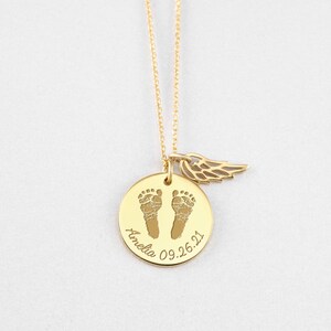Puede incluir: Un collar dorado con un colgante redondo que presenta dos huellas de beb&eacute; y el nombre "Amelia" con la fecha "09.26.21". Un peque&ntilde;o dije de ala cuelga junto al colgante. El collar est&aacute; sobre un fondo blanco.